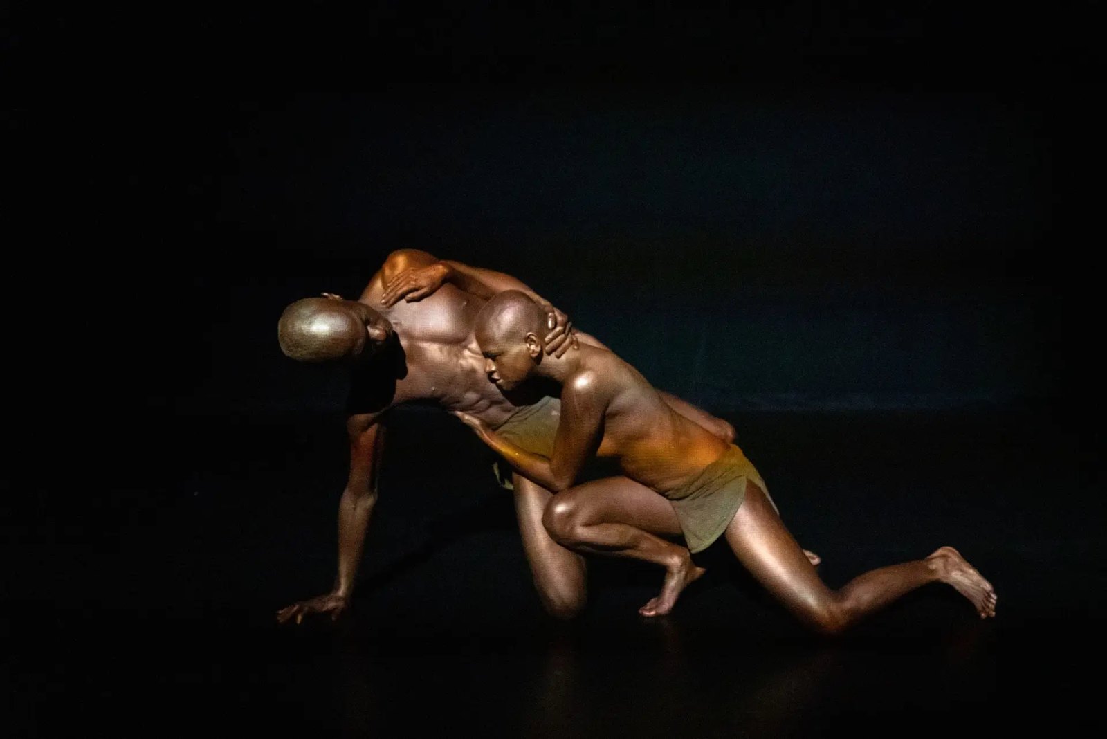 Two bronze-lit performers locked in a low crawling duet on a dark stage