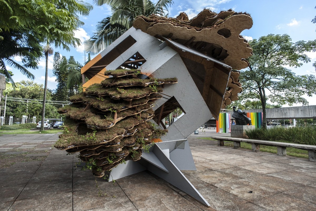 Tilted cube pavilion clad in layered earth and vegetation on the museum forecourt