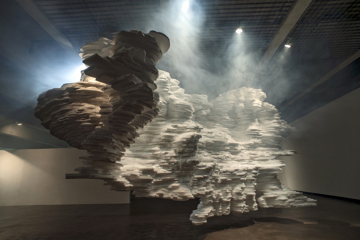 In Void — a suspended cloud of stacked white paper sheets filling a darkened gallery, lit through haze